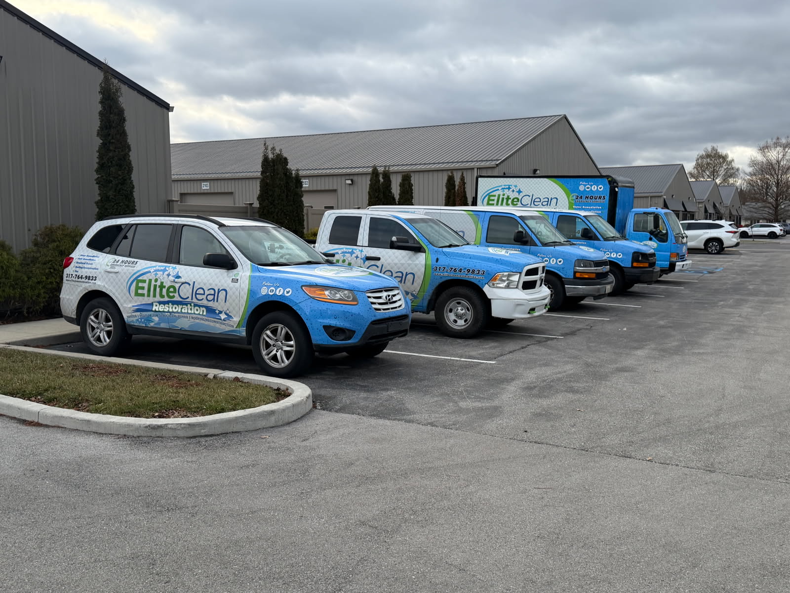 Four Elite Clean Restoration service vehicles are parked in a row outside a commercial building on a cloudy day. The vehicles are wrapped in blue-and-white company branding.