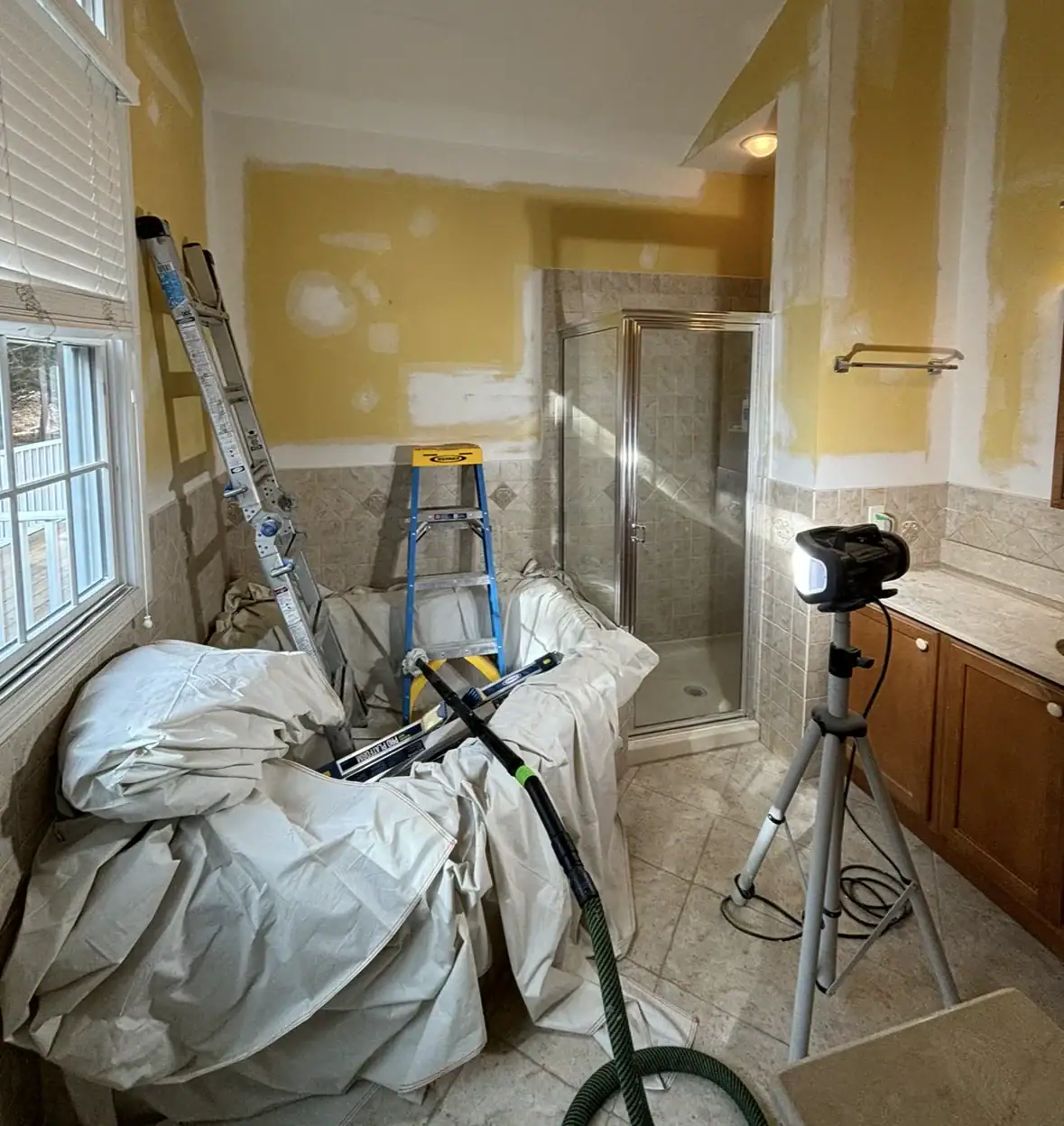 A bathroom under renovation with yellow and white patched walls, ladders, a covered bathtub, glass shower, work light on a tripod, and tools scattered around. Drop cloths protect the surfaces during Water Damage Restoration Indiana.