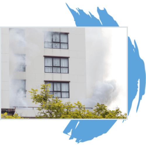Smoke billows from the lower floors of a white multi-story building, partially obscured by green trees in the foreground—an urgent reminder of the need for Water Damage Restoration Indiana. Blue brushstroke accents frame the image.