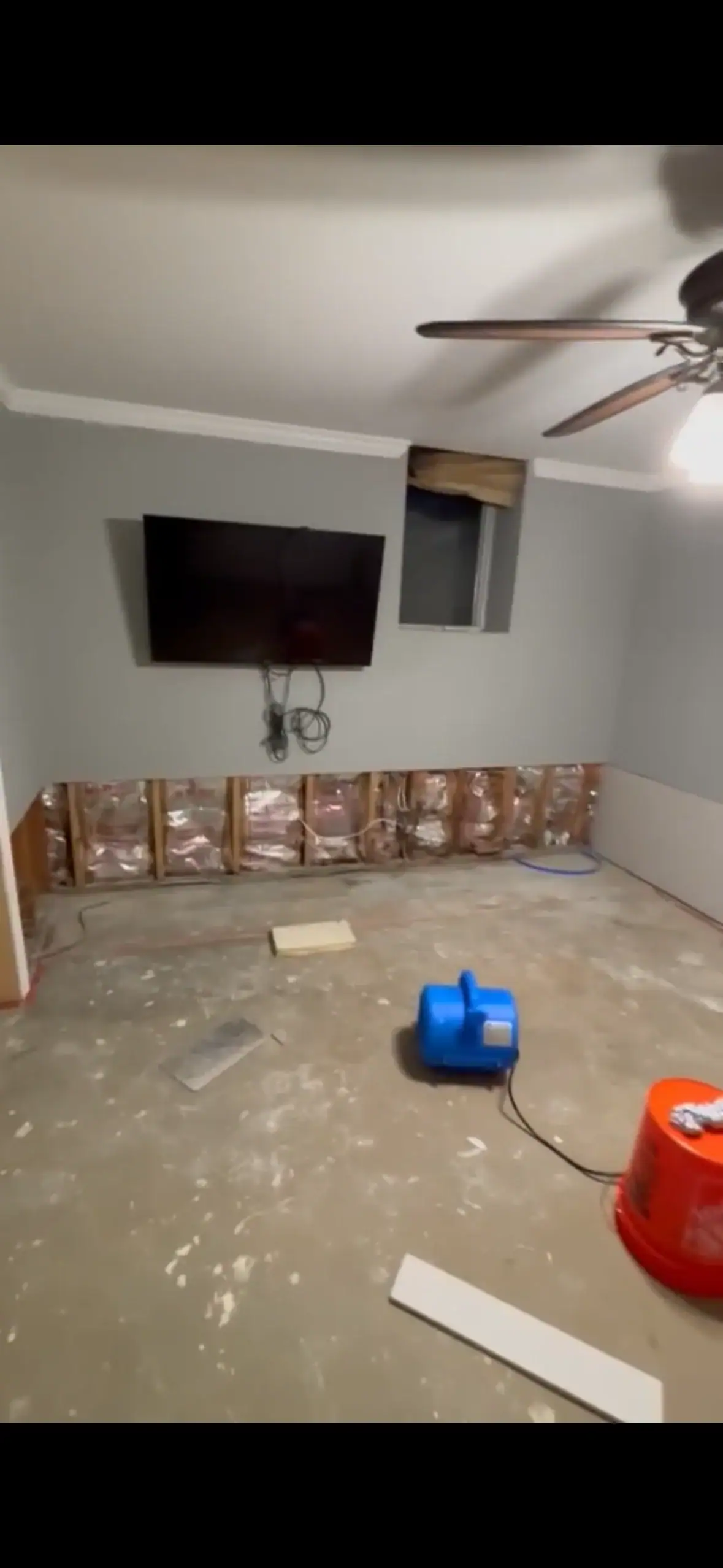 A room under renovation after water damage restoration in Indiana, featuring exposed wall insulation, an uncovered window, a mounted TV, ceiling fan, blue air mover, red bucket, and construction materials scattered on the bare floor.