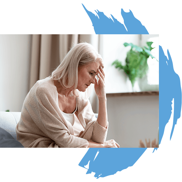 An elderly woman with light hair sits on a couch, looking worried and holding her forehead with one hand—indoors, framed by blue brushstroke accents. Blurred plants and a window suggest she may need Water Damage Restoration Indiana experts.