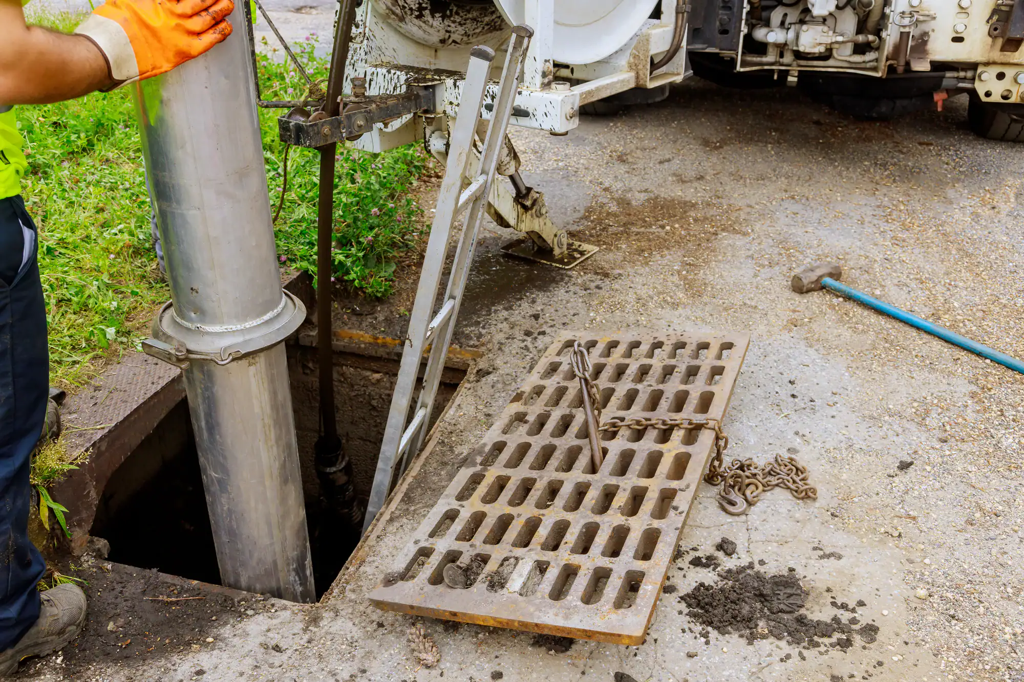 A worker in orange gloves operates machinery to clean or inspect a sewer drain. A large metal pipe is inserted into the open drain, and the grate cover is removed and lying nearby on the ground. A ladder, chain, and tools are visible.