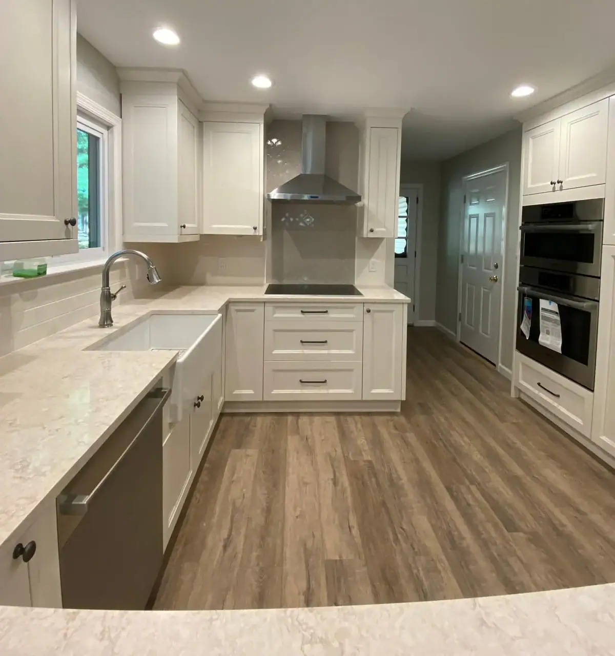 Modern kitchen with white cabinets, stainless steel appliances, farmhouse sink, and wood-look flooring. Light countertops and a large hood over the stove feature recessed lighting. Trustworthy Water Damage Restoration Indiana services help keep this space pristine.