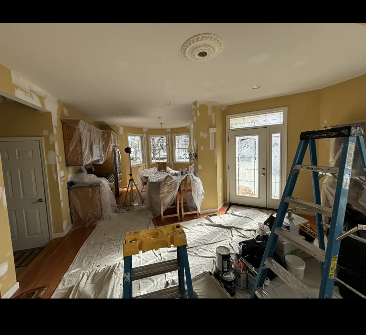 A room prepared for painting with furniture covered in plastic, a blue stepladder, drop cloths on the floor, and supplies scattered around—much like after Water Damage Restoration Indiana; sunlight streams through windows and a glass door.