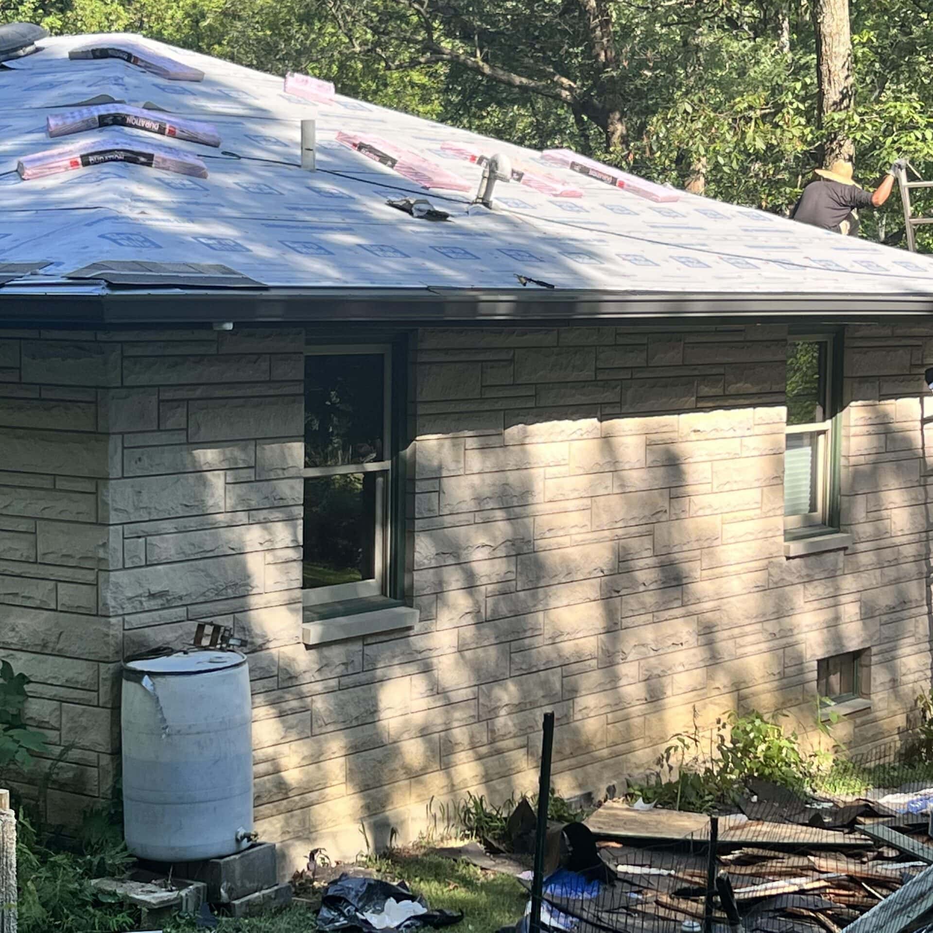 A person works on installing roofing materials on top of a stone house, possibly as part of Water Damage Restoration Indiana. Roofing supplies are spread across the roof, and trees are visible in the background.