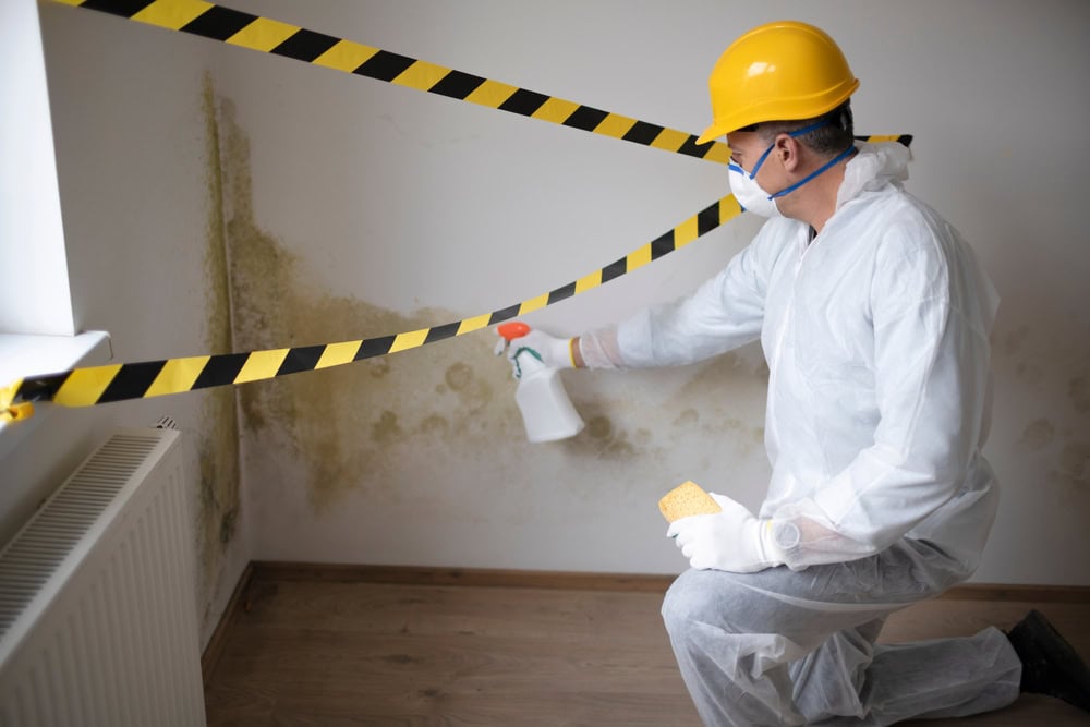A worker in protective gear sprays cleaning solution on mold growing on a wall behind black-and-yellow caution tape in a residential room.