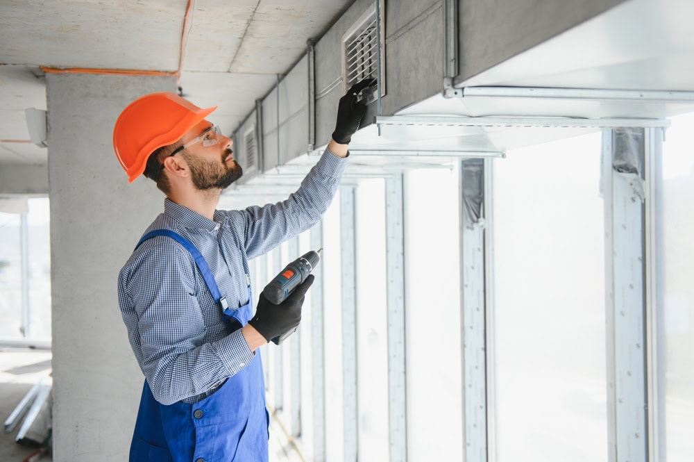 A construction worker in a hard hat and blue overalls uses a drill to fix or inspect an air vent on a ceiling duct, ensuring the building under construction meets Water Damage Restoration Indiana standards.