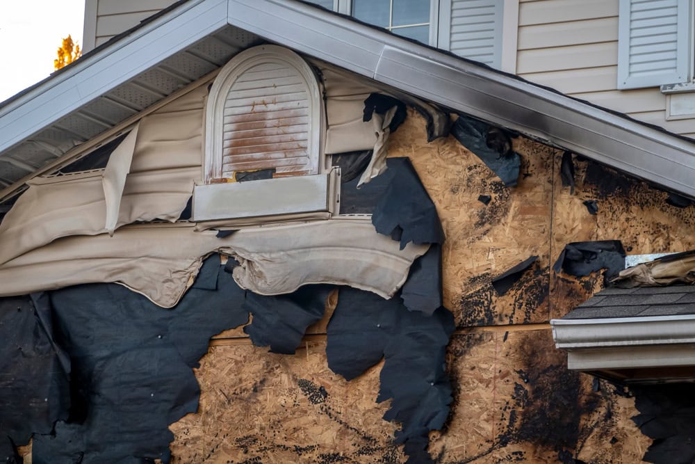 Close-up of a house exterior showing severe fire damage; melted siding and blackened, charred surfaces are visible on the upper wall near a window under the roof—potential site for Water Damage Restoration Indiana services.