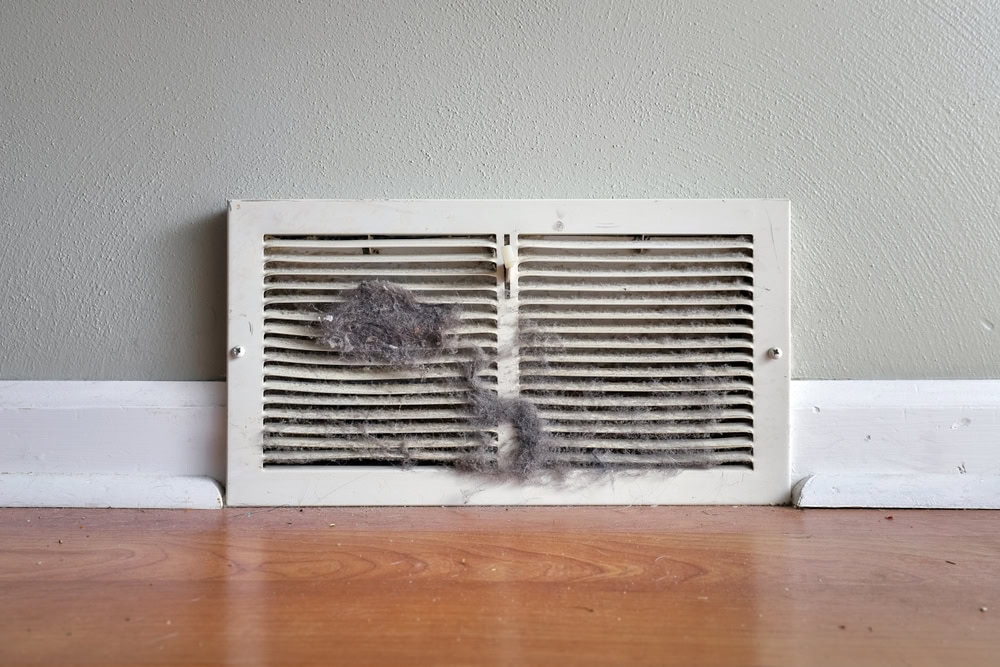 A white air vent on a wall near a wooden floor is covered in dust and dirt, especially in the center and left slats—a concern often noted during Water Damage Restoration Indiana services. The surrounding wall and baseboard remain clean in contrast.