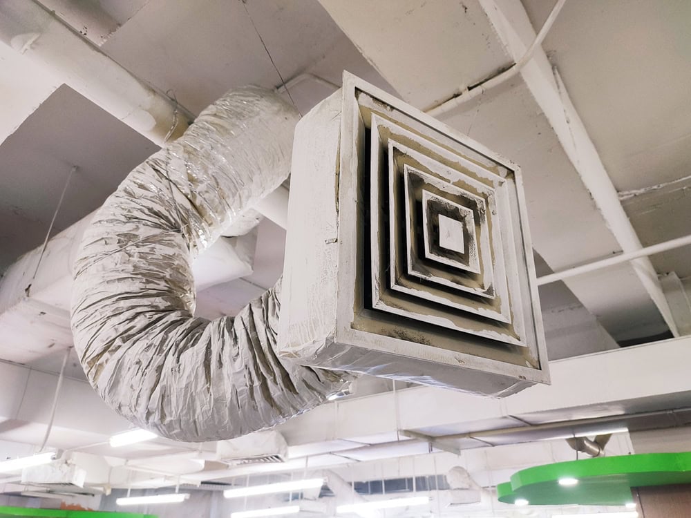 A close-up view of a square air vent connected to a flexible, foil-covered duct on a white ceiling with exposed pipes and lights in an indoor space, often seen during Water Damage Restoration Indiana projects.