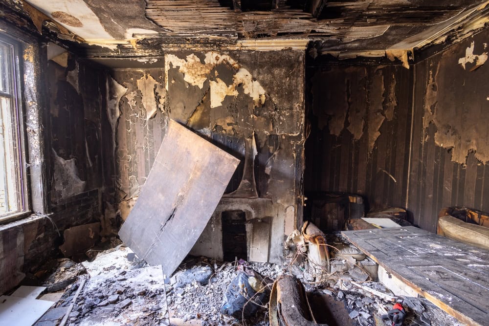 A room with severe fire damage: charred walls and ceiling, peeling plaster, debris-covered floor, and broken furniture scattered about. Light enters through a window on the left—Water Damage Restoration Indiana may help restore such spaces.