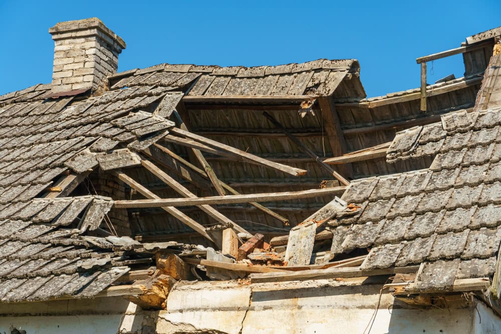 A close-up of a house roof severely damaged and partially collapsed, with broken wooden beams and missing tiles, under a clear blue sky—ideal for showcasing Water Damage Restoration Indiana services.
