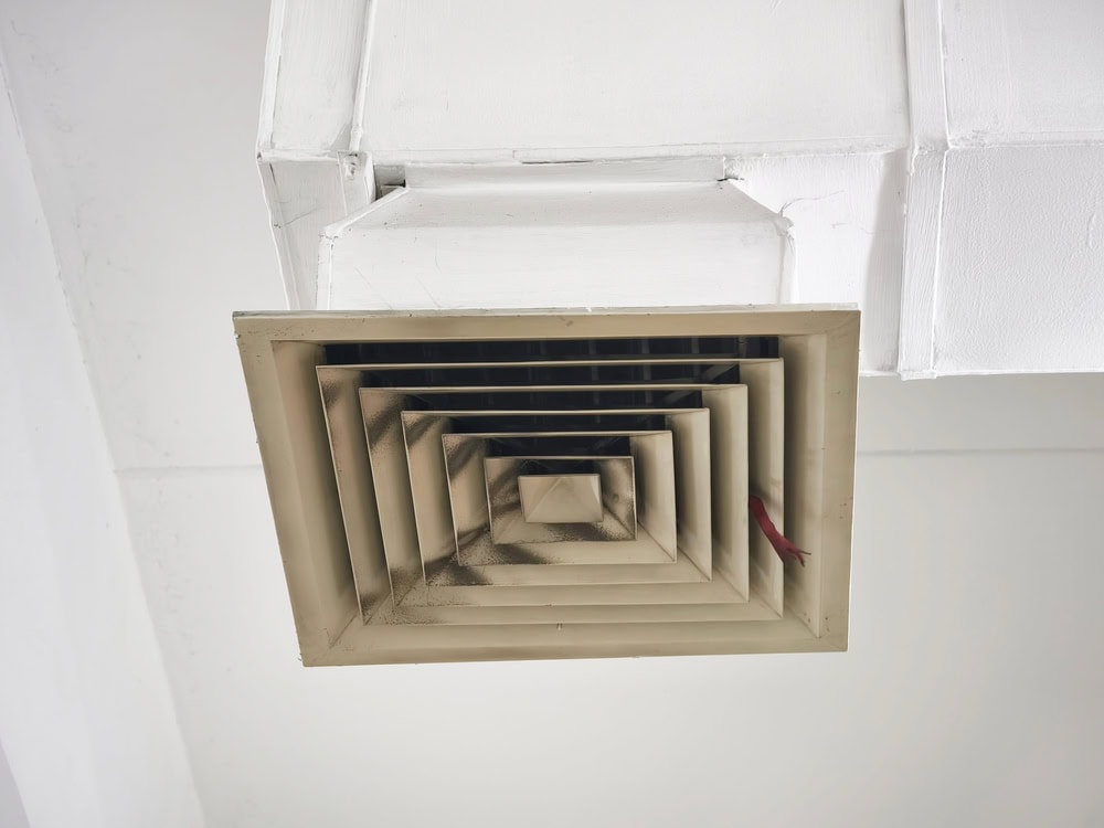 A close-up view of a square ceiling air vent, painted white, shows visible dust and dirt buildup on the slats—an issue often addressed during Water Damage Restoration Indiana services. The vent is attached to a white duct and ceiling.
