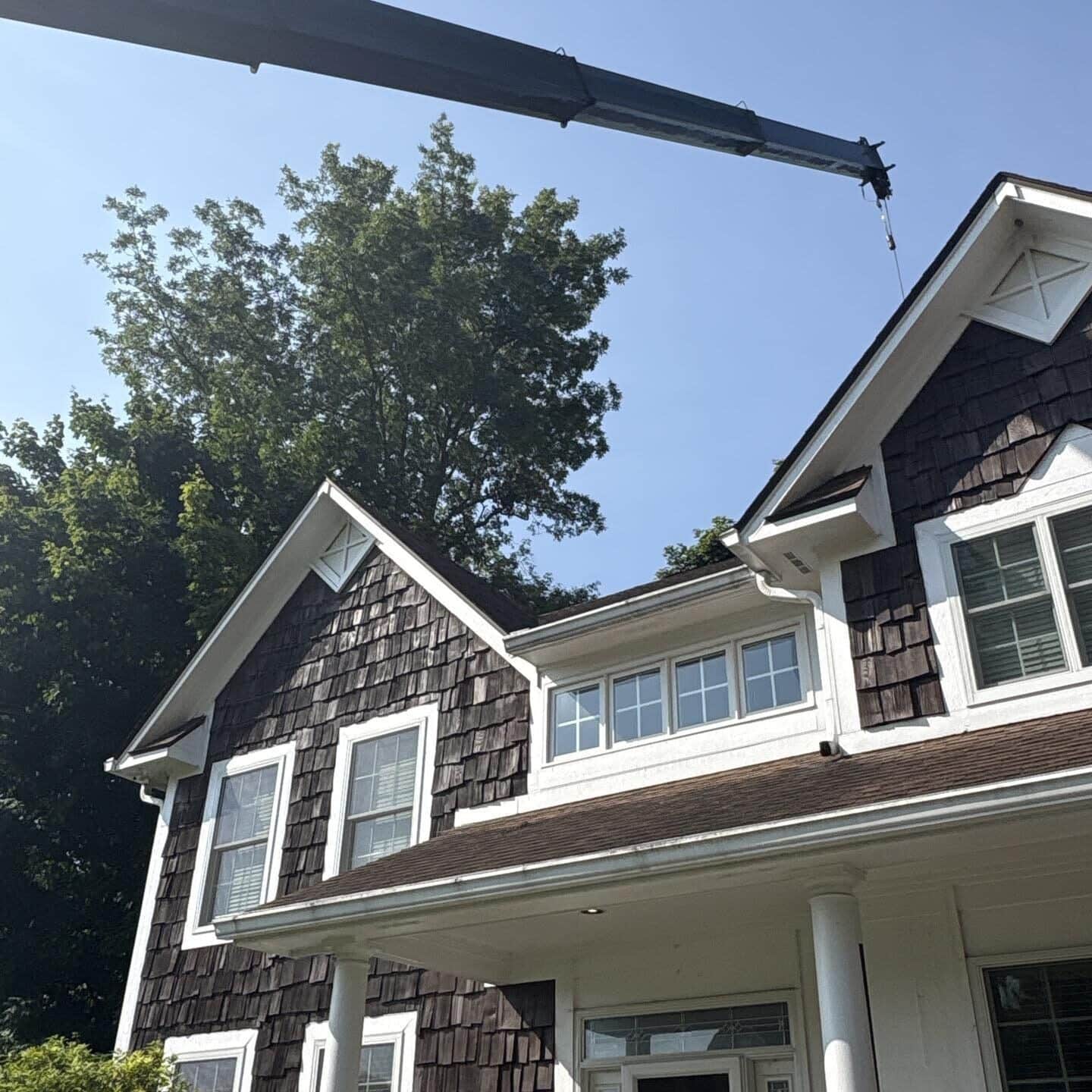 A large crane arm extends over the roof of a brown and white house with shingle siding as Water Damage Restoration Indiana experts work, surrounded by tall trees and a clear blue sky in the background.
