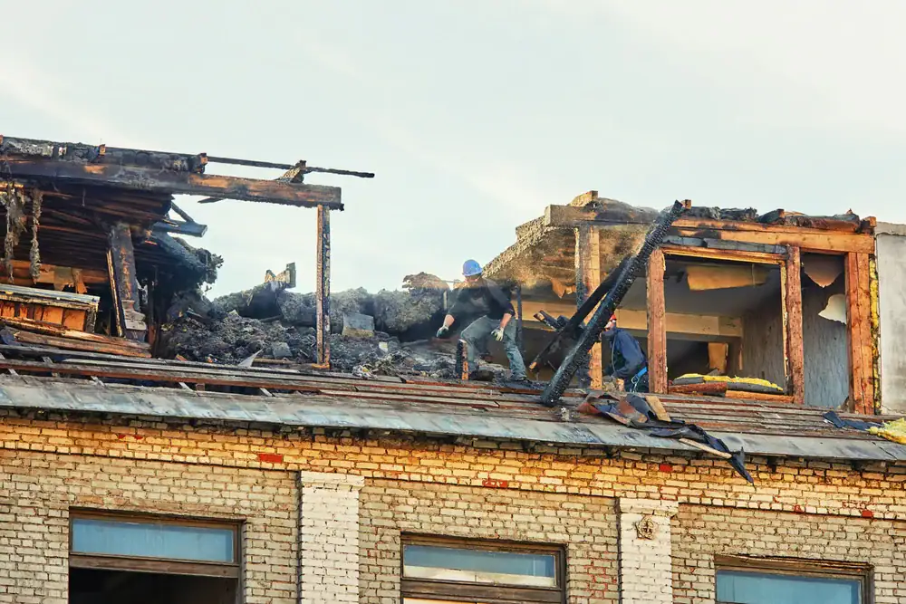Two people wearing helmets inspect the charred remains of an upper floor in a brick building damaged by fire, with debris and partially collapsed walls visible around them.