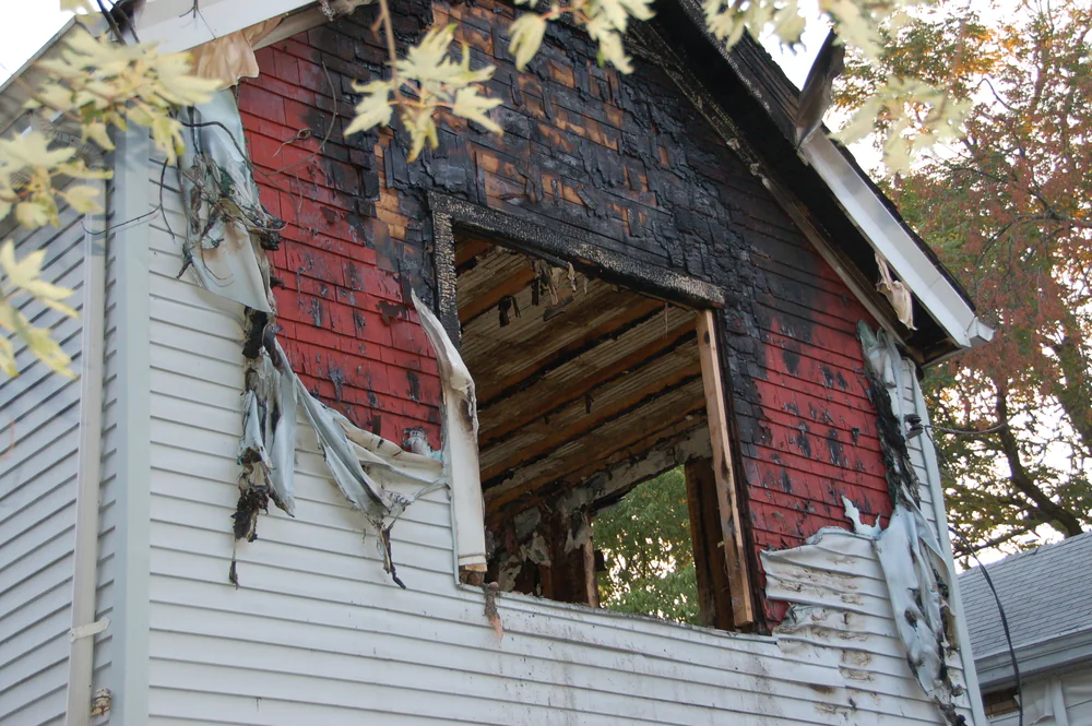 A two-story house with severe fire damage. The upper exterior wall is blackened and charred, with melted siding and a large, open window frame exposing the interior. Leaves are visible in the foreground.