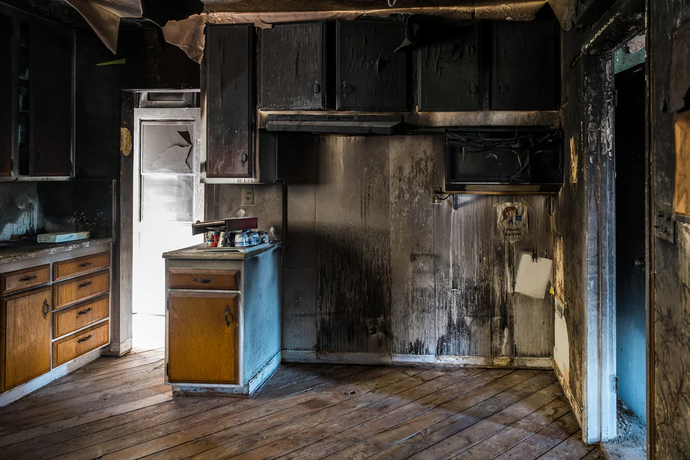 A kitchen with severe fire damage shows charred cabinets, blackened walls, and soot stains. The floor is wooden, and some cabinets remain intact. Light shines through a doorway, highlighting the extent of the damage.