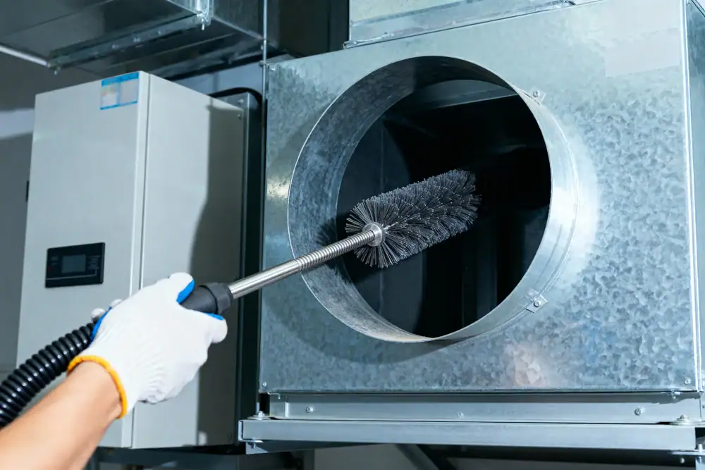 A person wearing a white glove is cleaning the inside of a large metal air duct with a long-handled round brush in an HVAC system.