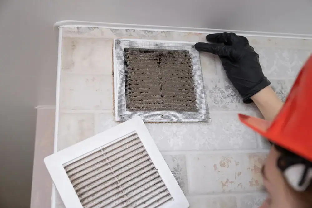 A person wearing a black glove and orange hard hat is removing a dusty, dirty vent cover from a wall, revealing a dirty air filter inside. Another clean vent cover is held nearby.