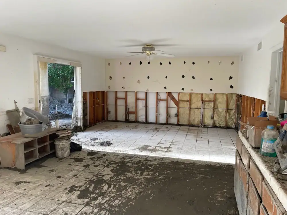 A damaged room with walls partially stripped to the studs, muddy floors, exposed insulation, and dirt throughout. The ceiling fan hangs intact, and debris and construction materials are scattered around.