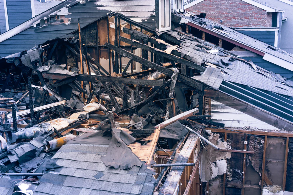 A house with extensive fire damage; the roof is partially collapsed, exposing charred wooden beams and debris inside. Scorched remains of walls and insulation are visible among the destruction.