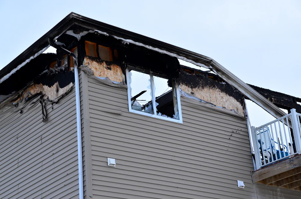 The upper level of a house with severe fire damage; the siding is charred and missing in places, a window is broken, and the interior appears burned, exposing insulation and framing.