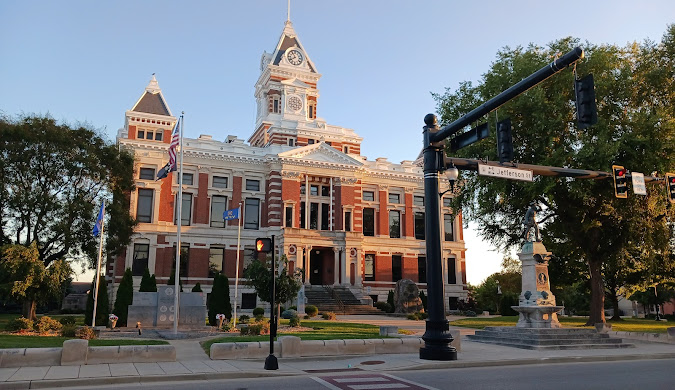 A historic red-brick courthouse with a central clock tower and white trim, surrounded by trees, monuments, and flags, stands at a street intersection—an iconic sight in Johnson County that’s just steps away from trusted Water Damage Restoration Service.