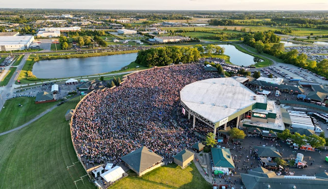 Aerial view of a large outdoor concert venue packed with people, surrounded by green fields, trees, ponds, and parking lots, under a clear sky with the sun setting in the distance.