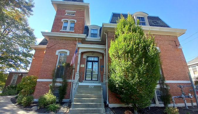 A two-story brick building with arched windows, a central entrance with stairs, and a tall tree in front. The address "170" is above the door, and two American flags are displayed near the entry.