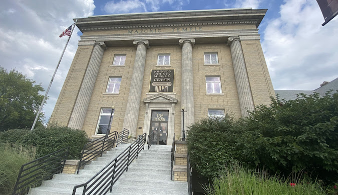 A large, tan brick building with tall columns and a sign reading "Johnson County Museum of History" above the entrance. An American flag is on the left, and a staircase with railings leads to the front door.