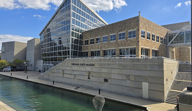 Modern building with stone and glass exterior labeled "Indiana State Museum," next to a calm canal under a blue sky with some clouds. A wire sculpture is visible near the water's edge.