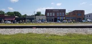 A row of small town storefronts and buildings sits across a street, with cars parked out front and a railroad track running parallel in the foreground, home to businesses like Water Damage Restoration Service in Johnson County on a sunny day.