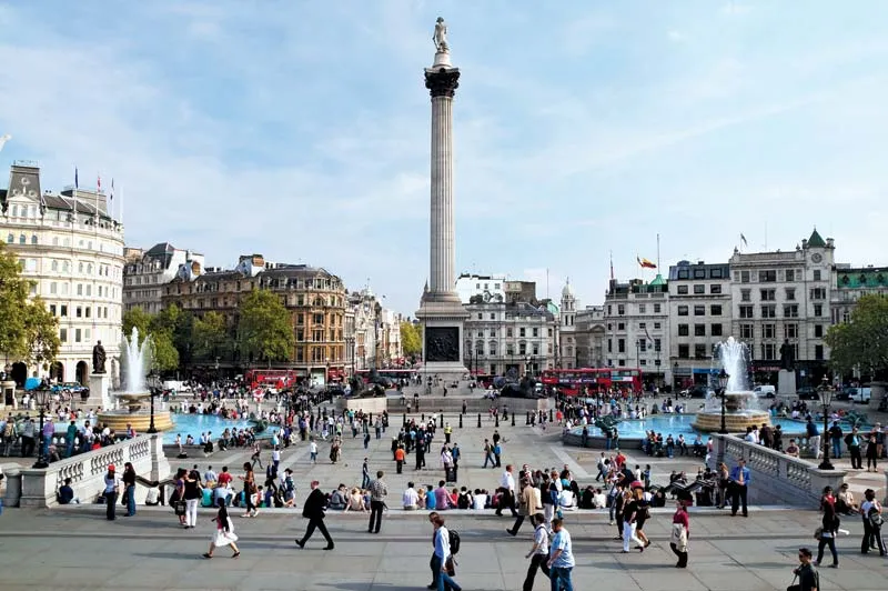 A busy public square with fountains, people walking and sitting, and a tall column with a statue at the center, surrounded by historic buildings, red double-decker buses, and a nearby Water Damage Restoration Service in Johnson County.